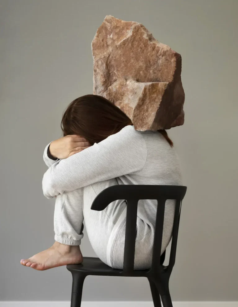 Mulher sentada encurvada em uma cadeira, com roupa de dormir, demonstrando cansaço fisico e emocional. Uma pedra grande e pesada está em suas costas para demostrar a importância de fazer a psicoterapia para depressão e se livrar deste peso.